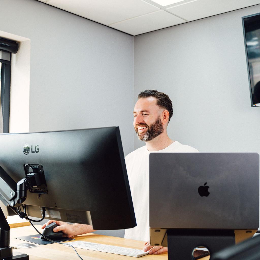 Een lachende man in een wit T-shirt staand achter een bureau. Op het bureau staat een scherm en een laptop.