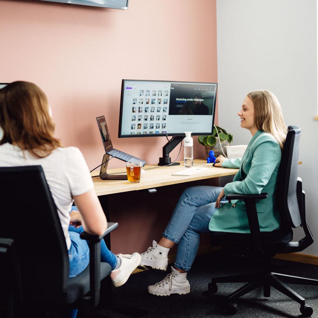 2 vrouwen van achter gefotografeerd, zittend aan hun bureau, met 2 schermen een laptop en een kopje thee.