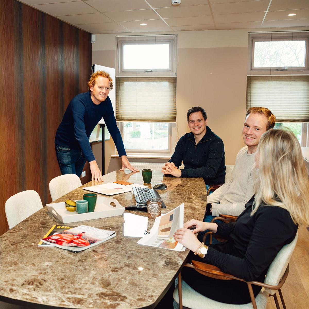 Een team van vier collega's in een vergaderruimte, waarbij één persoon presenteert bij een groot scherm met grafieken en data, terwijl de anderen luisteren.