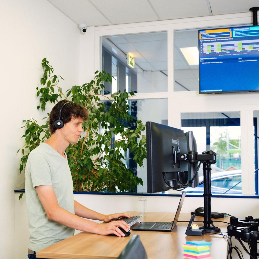 Een man in een lichtgroen shirt, staand achter zijn bureau met een tv scherm met belangrijke data op de achtergrond.