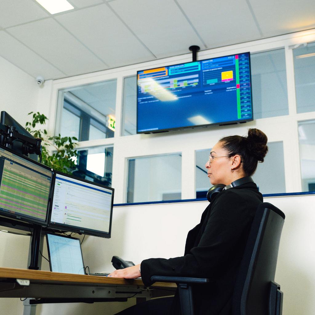 Een vrouw achter haar bureau met 2 grote schermen en een laptop. Een groot tv scherm met data op de achtergrond.