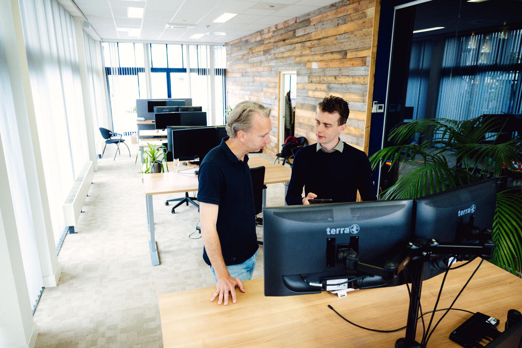 Twee mannen staan aan een bureau met daarop monitors met elkaar te praten.