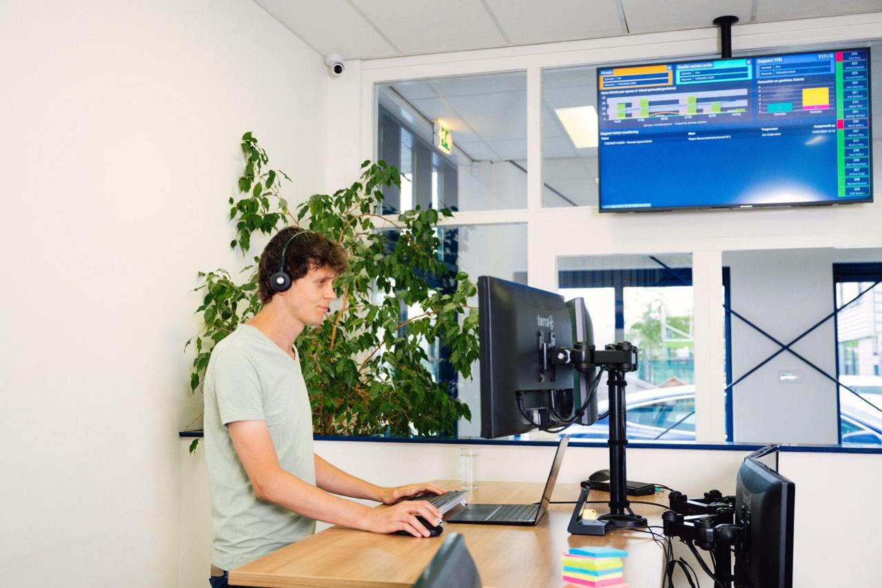 Man met koptelefoon op kijkt naar laptop en staat aan houten bureau met monitor boven hem die informatie toont. 