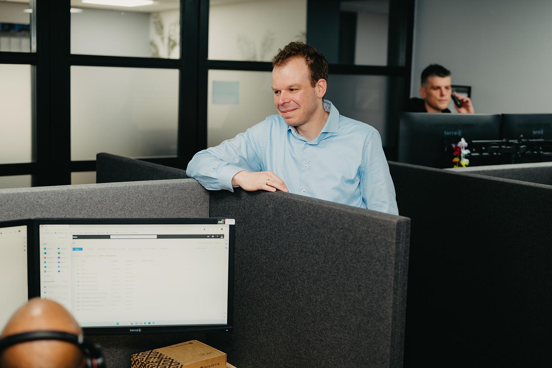 Een man bij een bureau kijkend naar een andere medewerker met monitor zichtbaar die informatie toont.