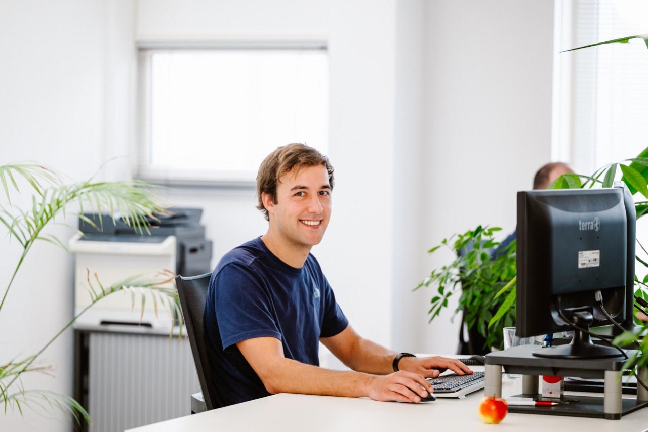 Man aan wit bureau met monitor en toetsenbord voor zich en glimlacht naar de camera.