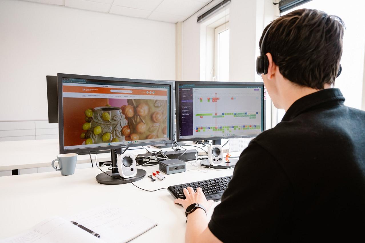 Blik op twee monitoren op een wit bureau meekijkend over de schouder van een man met koptelefoon op.
