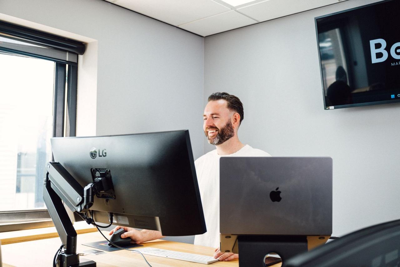 Man die glimlacht staat achter bureau met monitor en laptop voor zich.