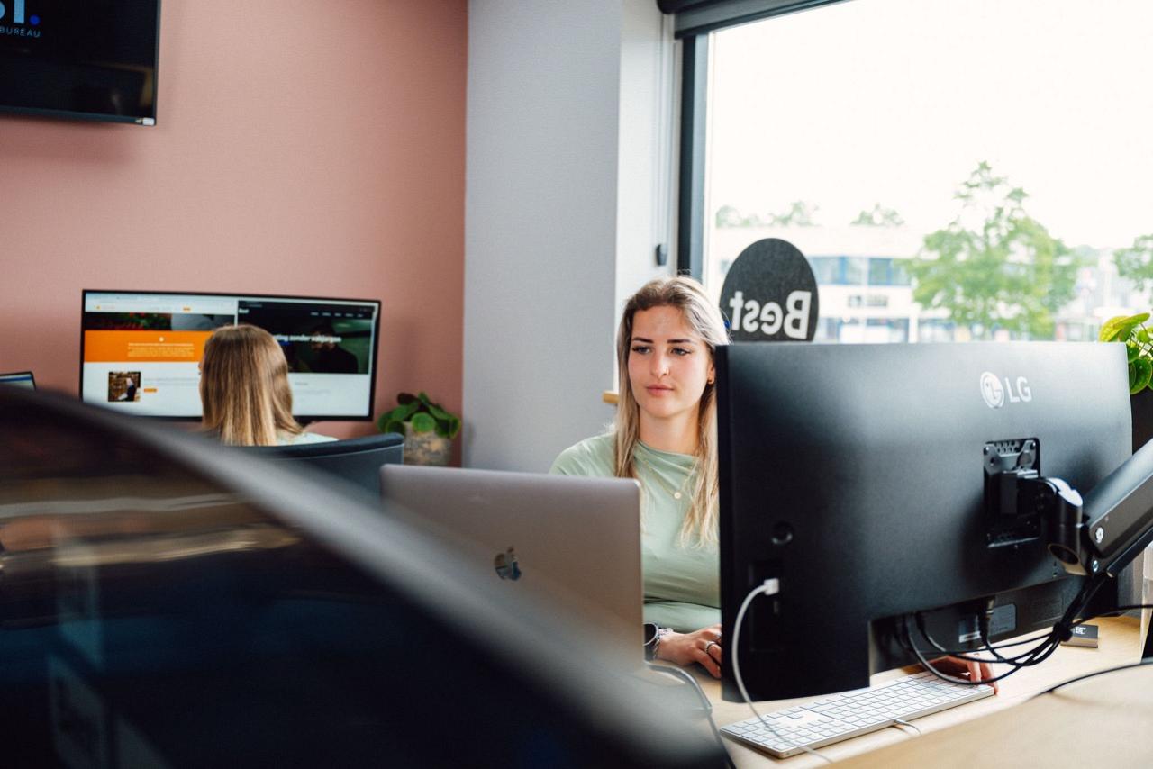 Kantooromgeving met monitoren en vrouw achter een bureau kijkend op haar scherm.