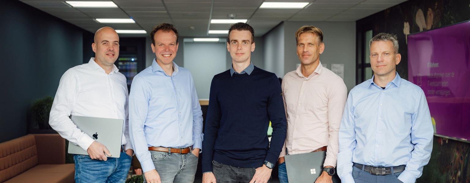 Een groep van vijf mannen poseert in een modern kantoor. Ze dragen zakelijke kleding en houden laptops vast. De sfeer straalt professionaliteit uit.