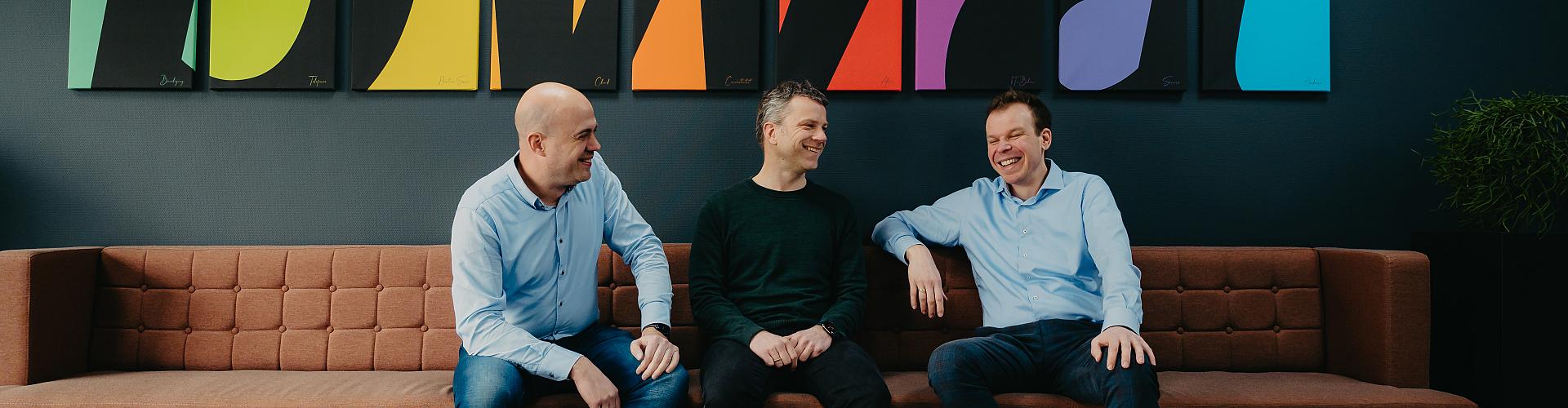 Drie mannen op een bruine bank voor een kleurig PWA-logo lachen ergens om.