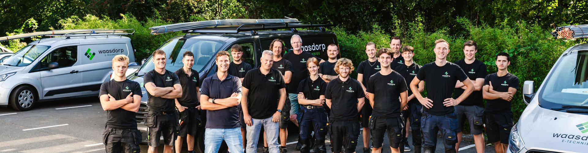 Groep mannen in zwarte shirts staan op parkeerplaats tussen auto's.