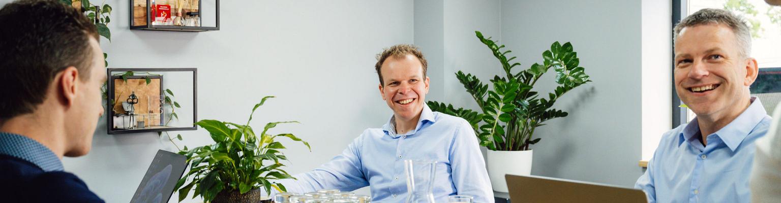 Vier collega’s in een overleg in een moderne vergaderruimte, met laptops, waterglazen en planten, in een ontspannen en professionele sfeer.