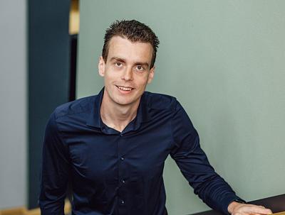Man in een donkerblauw overhemd kijkt met een open glimlach in de camera. Hij leunt op een oppervlak, met een rustige, neutrale achtergrond.