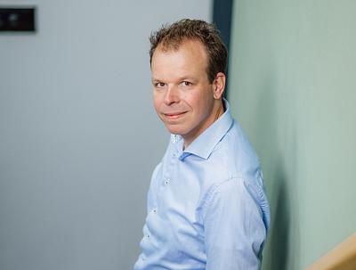 Man in een lichtblauw overhemd leunt ontspannen tegen een muur en kijkt vriendelijk in de camera. De achtergrond is neutraal en straalt rust uit.