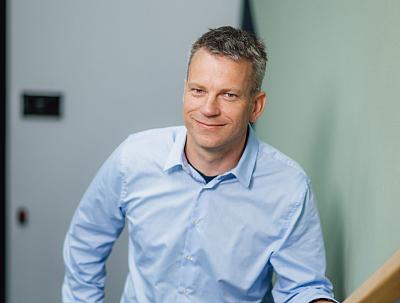 Man in lichtblauw overhemd glimlacht terwijl hij leunt tegen een trapleuning in een moderne kantooromgeving met een rustige uitstraling.