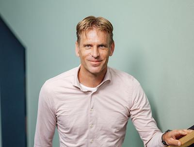 Man in een lichtroze overhemd glimlacht zelfverzekerd naar de camera. Hij leunt lichtjes naar voren, met een neutrale achtergrond op de achtergrond.