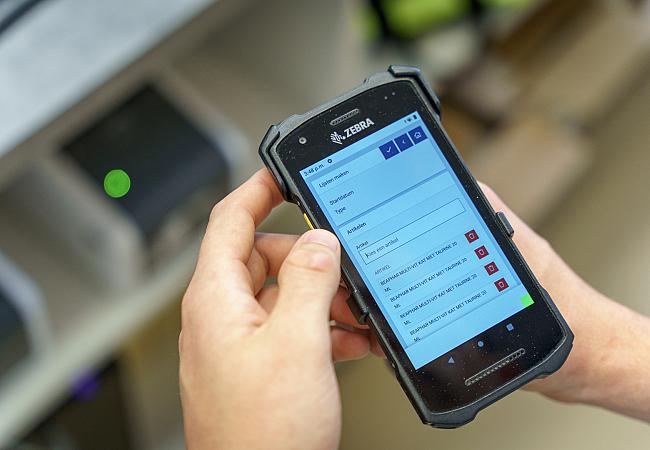 Hand toont handscanner met daarop informatie met op de achtergrond een kassa.